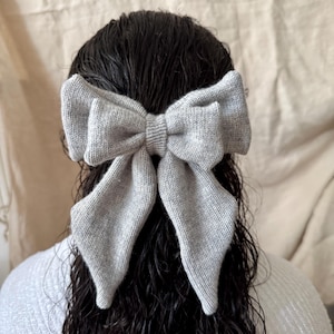May include: A large, light gray knitted bow hair accessory. The bow is centered on dark, wet hair. The bow is made of a soft, textured material and has long, flowing tails. The hair accessory is a stylish addition to any hairstyle.