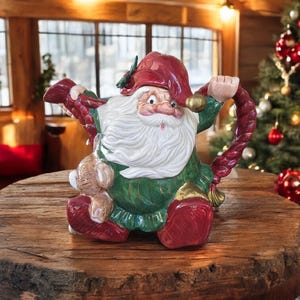 May include: A ceramic Santa Claus teapot with a red hat and boots, green coat, and white beard. The teapot has a braided handle and holds a small teddy bear. The teapot is on a wooden table.