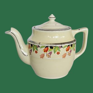 May include: Cream-colored ceramic teapot with a lid and handle. The teapot features a floral design with red, orange, purple, and green accents. A silver trim accents the rim and spout. The teapot is set against a green background.