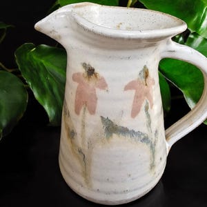 May include: A handmade ceramic pitcher with a white base and a curved handle. The pitcher features two pink flower designs and green and brown accents. The pitcher has a spout for easy pouring and is suitable for serving beverages.