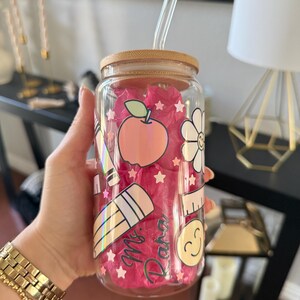 May include: A clear glass tumbler with a bamboo lid and a glass straw. The tumbler is decorated with school-themed stickers, including an apple, a flower, a pencil, and the name "Mr. Dana". The tumbler is filled with pink tissue paper.