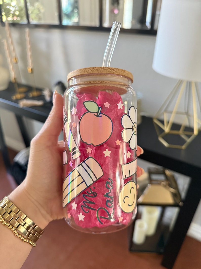 May include: A clear glass tumbler with a bamboo lid and a glass straw. The tumbler is decorated with school-themed stickers, including an apple, a flower, a pencil, and the name "Mr. Dana". The tumbler is filled with pink tissue paper.