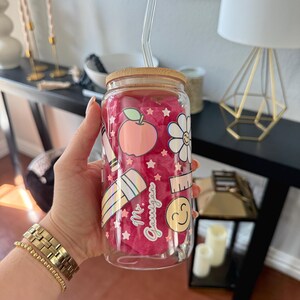 May include: A clear glass tumbler with a bamboo lid and a glass straw. The tumbler features a pink interior and is decorated with school-themed graphics, including an apple, pencil, and ruler. The text "Mr. Gauigan" is also visible on the tumbler.