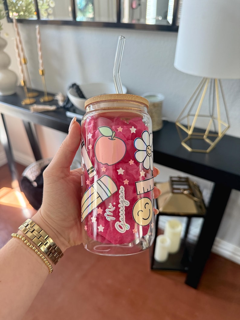 May include: A clear glass tumbler with a bamboo lid and a glass straw. The tumbler features a pink interior and is decorated with school-themed graphics, including an apple, pencil, and ruler. The text "Mr. Gauigan" is also visible on the tumbler.