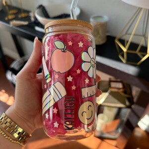 May include: A clear glass tumbler with a bamboo lid and a clear straw. The tumbler is decorated with school-themed holographic stickers, including an apple, flower, ruler, pencil, and a smiling face. The name "KALEIGH" is printed on the glass.