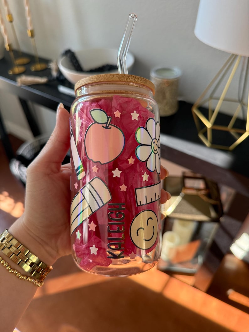 May include: A clear glass tumbler with a bamboo lid and a clear straw. The tumbler is decorated with school-themed holographic stickers, including an apple, flower, ruler, pencil, and a smiling face. The name "KALEIGH" is printed on the glass.
