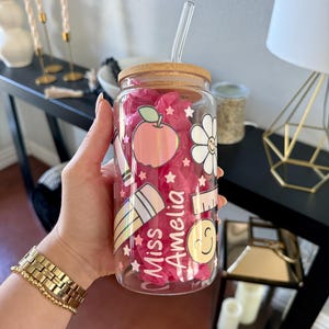 May include: A clear glass tumbler with a bamboo lid and a clear straw. The tumbler is decorated with school-themed graphics, including an apple, pencil, and a smiling face. The text "Miss Amelia" is also featured on the glass.
