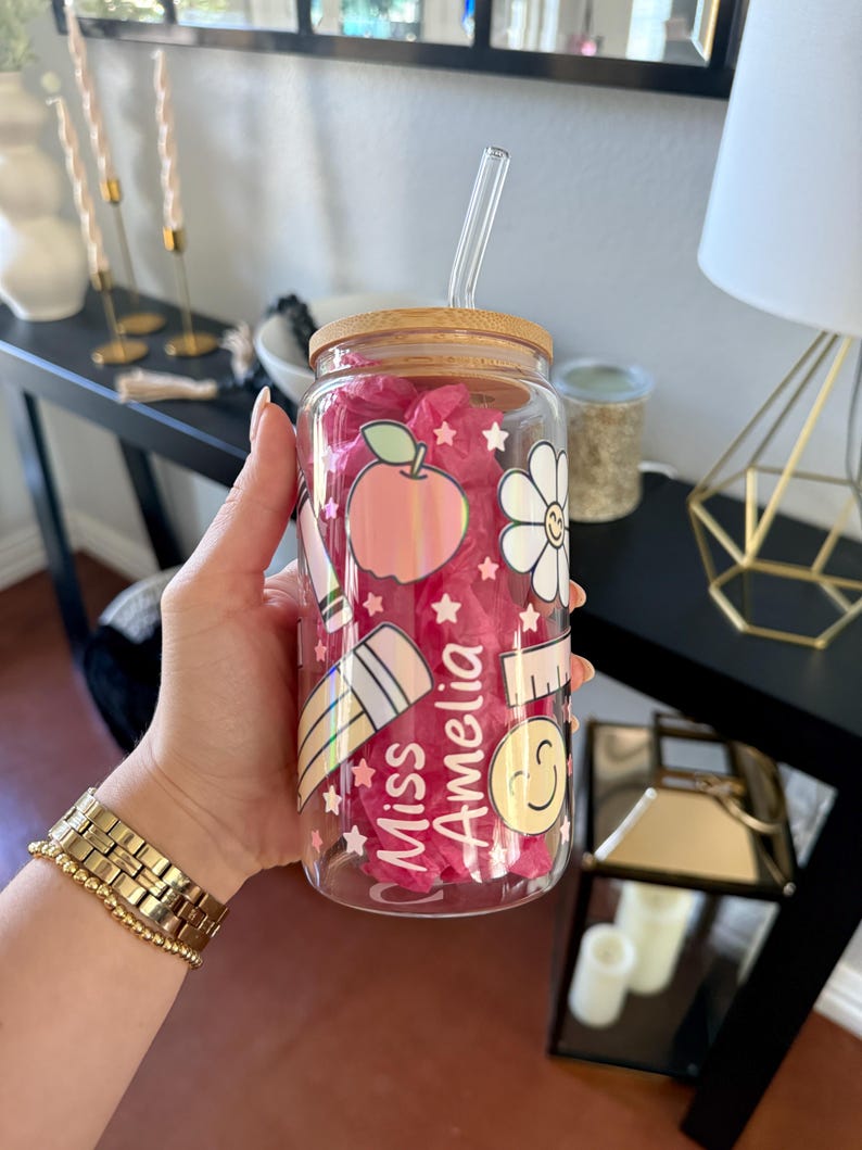 May include: A clear glass tumbler with a bamboo lid and a clear straw. The tumbler is decorated with school-themed graphics, including an apple, pencil, and a smiling face. The text "Miss Amelia" is also featured on the glass.