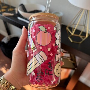 May include: A clear glass tumbler with a bamboo lid and a glass straw. The tumbler is decorated with school-themed stickers, including an apple, pencil, and ruler. The text "Ms. Flor" is also visible on the glass. The tumbler is filled with pink tissue paper.