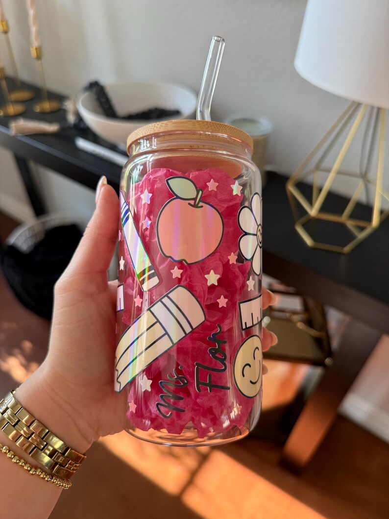 May include: A clear glass tumbler with a bamboo lid and a glass straw. The tumbler is decorated with school-themed stickers, including an apple, pencil, and ruler. The text "Ms. Flor" is also visible on the glass. The tumbler is filled with pink tissue paper.