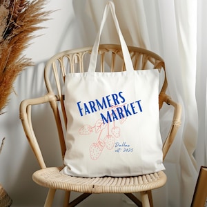 May include: A white tote bag with blue text that reads "FARMERS MARKET" and a pink illustration of strawberries. The bag has long handles and is sitting on a wicker chair. The text "Dallas est. 2025" is also on the bag.