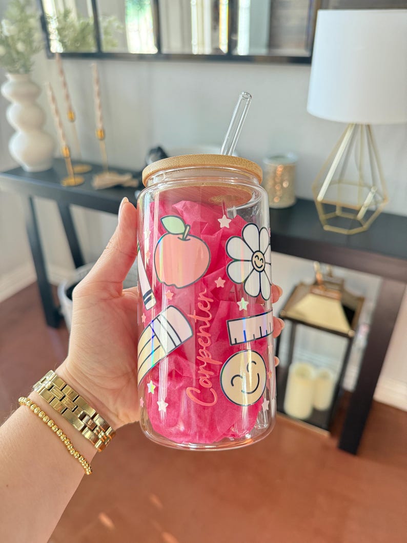 May include: A clear glass tumbler with a bamboo lid and a clear straw. The tumbler is decorated with colorful school-themed stickers, including an apple, a flower, and a ruler. The word "Carpenter" is written in pink script on the side.