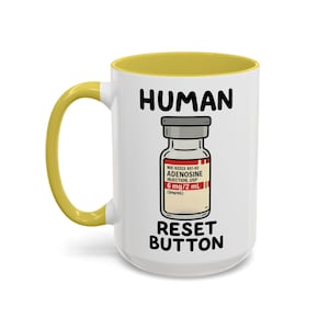 May include: A white ceramic mug with a yellow interior and handle. The mug features the words "HUMAN" and "RESET BUTTON" in black, with an illustration of an adenosine injection vial. The vial has red and white labels.