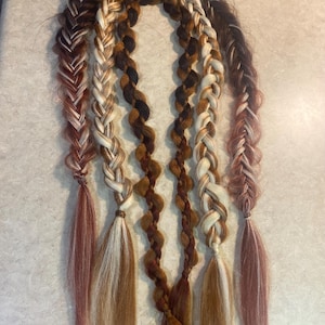 Op de afbeelding: Een verzameling gevlochten haarverlengingen in verschillende tinten bruin, blond en roodbruin. De vlechten zijn in een gebogen patroon gerangschikt en tonen verschillende vlechtstijlen en kleurencombinaties. De uiteinden van de vlechten zijn los en vloeiend.