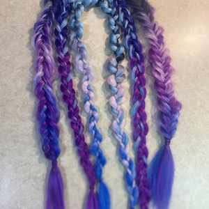 Puede incluir: Extensiones de cabello trenzadas en tonos morados, azules y negros. Las extensiones están dispuestas en un estilo de trenza de espiga, con los colores mezclándose de oscuro a claro. Los extremos de las trenzas son sueltos.