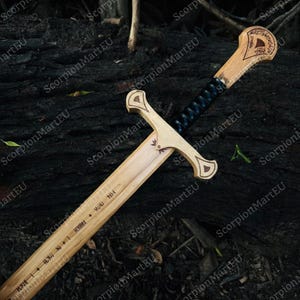 May include: A wooden sword with a light brown blade and handle, featuring a black wrapped grip. The handle has decorative carvings and text. The sword is displayed on a dark, textured surface.
