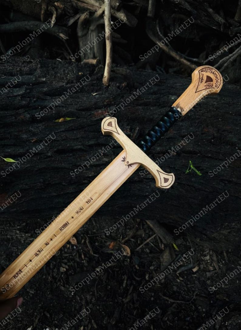 May include: A wooden sword with a light brown blade and handle, featuring a black wrapped grip. The handle has decorative carvings and text. The sword is displayed on a dark, textured surface.