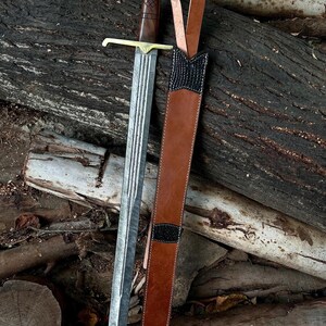 Peut inclure: Une épée de style médiéval avec une lame argentée et un manche en bois marron. L'épée a une garde transversale dorée et un pommeau décoratif. Un fourreau en cuir marron avec des accents noirs est à côté de l'épée.