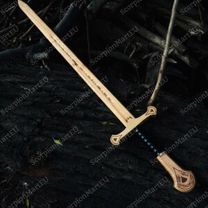 May include: A wooden sword with a light brown blade and handle, featuring a black grip and decorative carvings. The sword is laid on a dark, textured surface, possibly wood or bark. The blade has etched text.