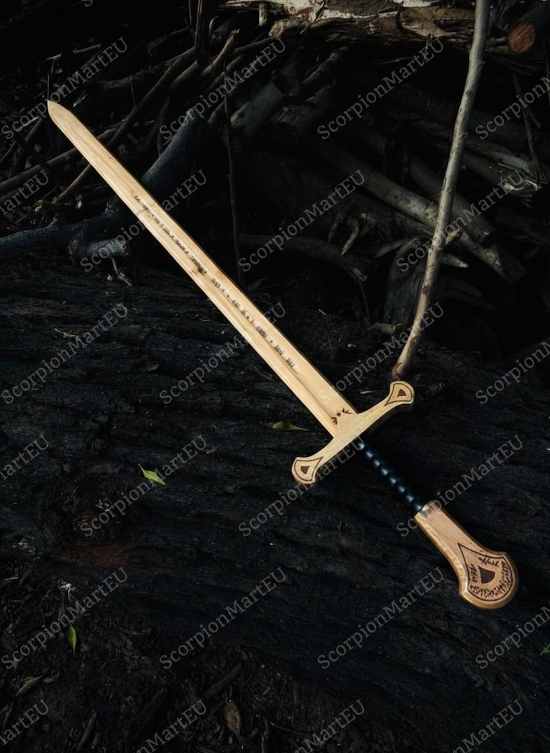 May include: A wooden sword with a light brown blade and handle, featuring a black grip and decorative carvings. The sword is laid on a dark, textured surface, possibly wood or bark. The blade has etched text.