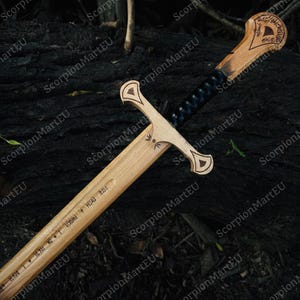 May include: A wooden sword with a light brown blade and handle, featuring intricate carvings and a dark blue wrapped grip. The blade has engraved text. The sword is displayed against a dark, textured background.