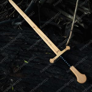 May include: A wooden sword with a light brown blade and a darker brown handle. The handle has a black grip and a crossguard. The sword is lying on a dark surface.