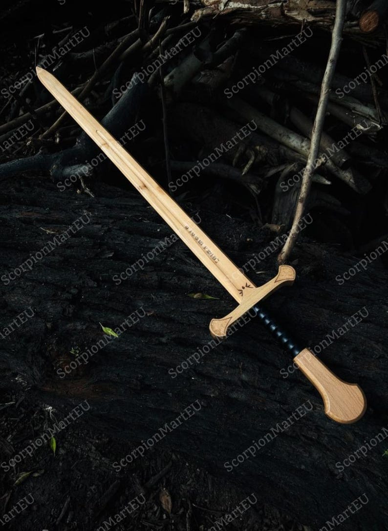 May include: A wooden sword with a light brown blade and a darker brown handle. The handle has a black grip and a crossguard. The sword is lying on a dark surface.