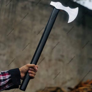 Könnte beinhalten: Eine Axt mit schwarzem Griff und silberner Klinge. Der Axtkopf hat ein geschwungenes Design. Der Griff ist lang und gerade. Die Axt wird in einer Hand gehalten, wobei ein karierter Hemdärmel sichtbar ist.