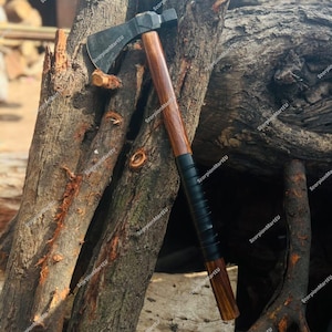 Könnte beinhalten: Axt mit einem dunklen Metallkopf und einem Holzgriff. Der Griff ist in der Nähe des Kopfes mit schwarzem Leder umwickelt. Die Axt lehnt an einem verwitterten Baumstamm. Die Axt ist ein Werkzeug zum Holzhacken.