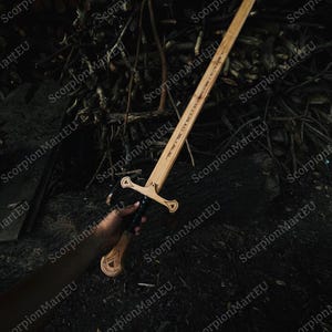 May include: A wooden sword with a light brown blade and a detailed handle. The sword has a crossguard and text etched along the blade. The handle is dark, and the sword is held against a dark background of twigs and leaves.