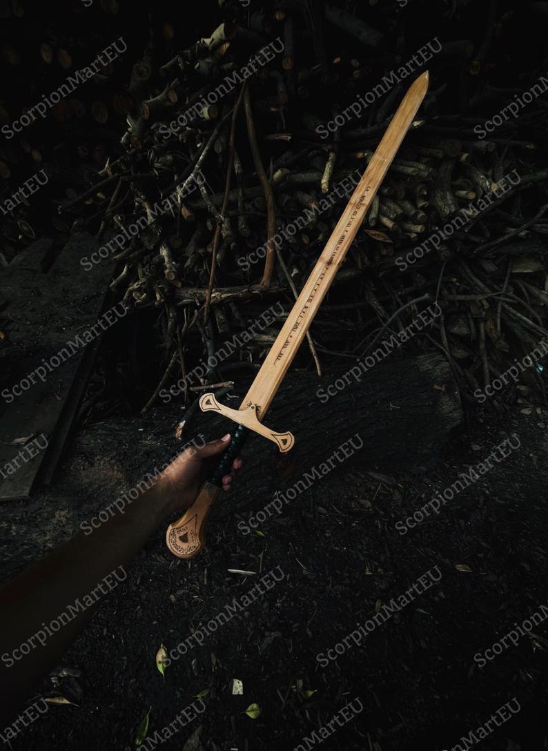 May include: A wooden sword with a light brown blade and a detailed handle. The sword has a crossguard and text etched along the blade. The handle is dark, and the sword is held against a dark background of twigs and leaves.