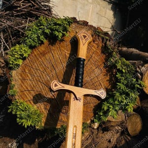 May include: A wooden sword with a light brown blade and handle, featuring a black grip. The handle has decorative carvings and the blade has engraved text. The sword is displayed on a wooden log with green foliage.