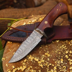 May include: A handcrafted hunting knife with a patterned steel blade and a dark wood handle. The knife is accompanied by a brown leather sheath. The blade has a unique wave-like pattern. The handle is adorned with brass accents.