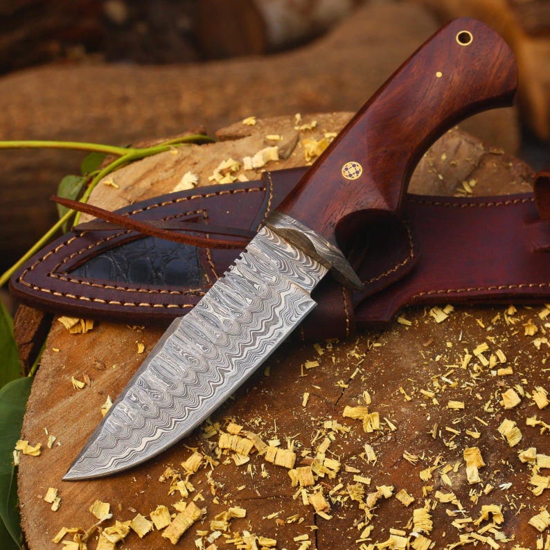 May include: A handcrafted hunting knife with a patterned steel blade and a dark wood handle. The knife is accompanied by a brown leather sheath. The blade has a unique wave-like pattern. The handle is adorned with brass accents.