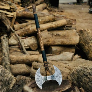 May include: A double-bladed axe with a wooden handle and silver-colored blades. The handle is intricately carved with a dark blue grip. The blades feature a Celtic knot design. The axe is displayed on a wooden surface.