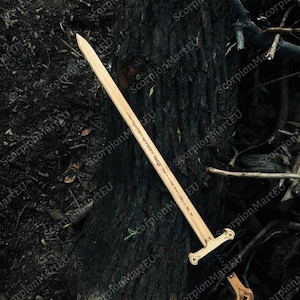 May include: A wooden sword with a light brown blade and a detailed handle. The sword is lying on a dark, textured surface, possibly a tree trunk. The handle has a decorative design.