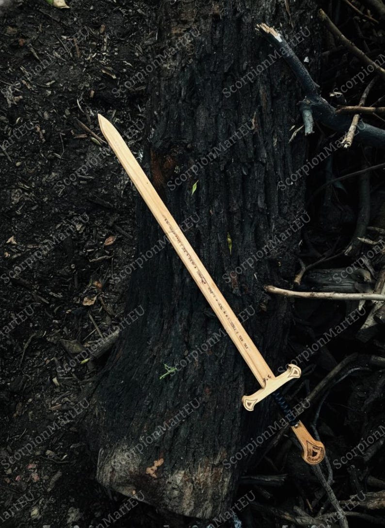 May include: A wooden sword with a light brown blade and a detailed handle. The sword is lying on a dark, textured surface, possibly a tree trunk. The handle has a decorative design.