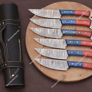 May include: Set of six Damascus steel knives with red and blue handles, displayed on a wooden cutting board. A black leather knife roll is on the left. The blades have a unique patterned design.