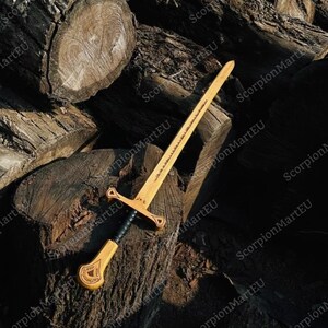 May include: A wooden sword with a light brown blade and a black handle, resting on a pile of cut logs. The sword features a crossguard and a detailed hilt design. The blade has etched text.
