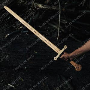 May include: A wooden sword with a light brown blade and a decorative handle. The sword is held by a person's hand against a dark background of wood and foliage. The sword has a cross-shaped hilt.