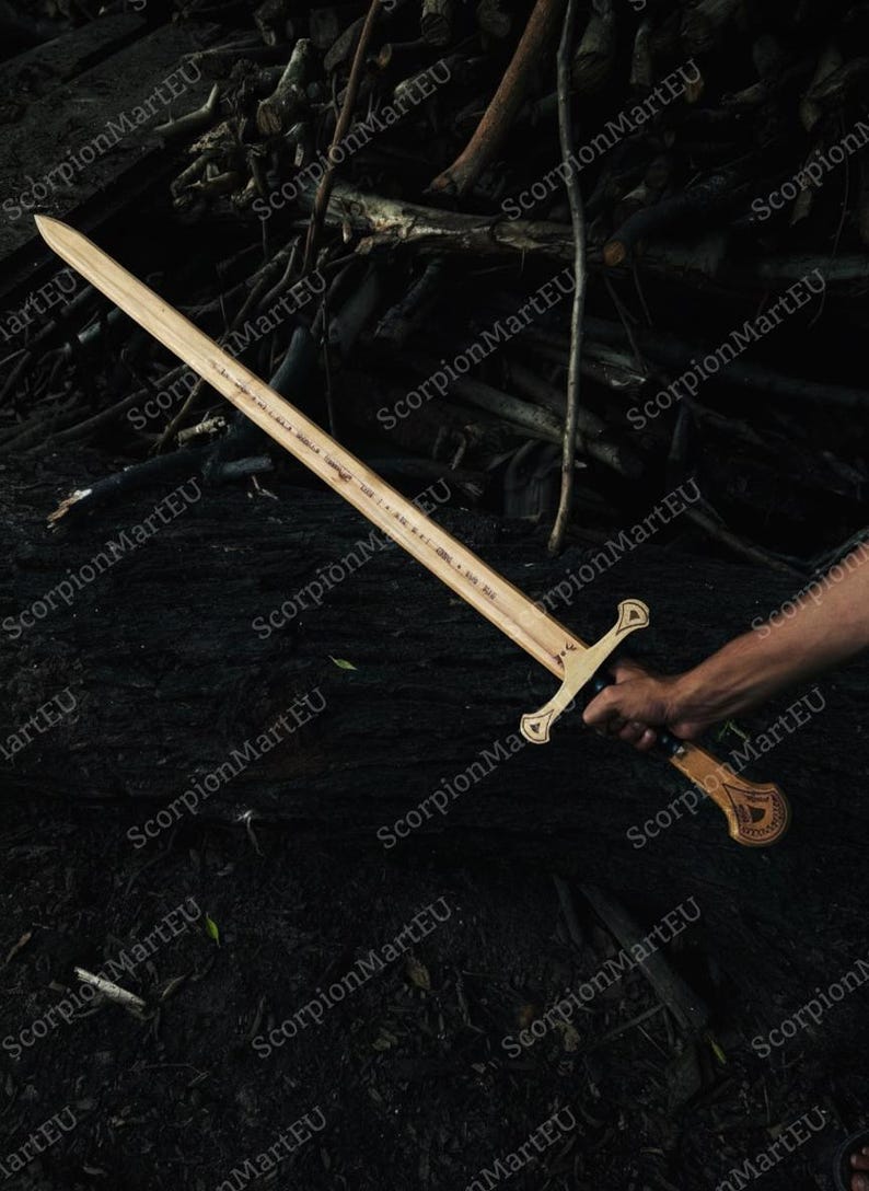 May include: A wooden sword with a light brown blade and a decorative handle. The sword is held by a person's hand against a dark background of wood and foliage. The sword has a cross-shaped hilt.