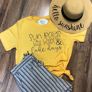 Op de afbeelding: Geel T-shirt met de tekst "Sun Rays Boat Waves & Lake Days" in zwart. Een strohoed met de tekst "hello sunshine" in zwart ligt op het T-shirt. Een gestreepte short ligt op het T-shirt.