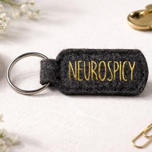 Könnte beinhalten: Ein dunkelgrauer Filz-Schlüsselanhänger mit dem Wort "NEUROSPICY" in goldfarbenem Faden gestickt. Der Schlüsselanhänger hat einen silbernen Metallring. Weitere Elemente im Bild sind goldene Büroklammern, Schlüssel und weiße Blumen.
