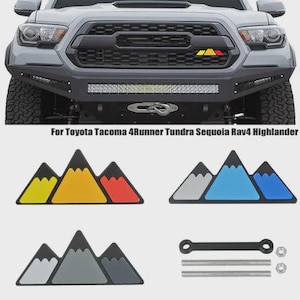Puede incluir: Un conjunto de emblemas para automóviles con forma de montaña en varias combinaciones de colores, incluyendo amarillo, naranja, rojo, gris y azul. Los emblemas se muestran con accesorios de montaje y están diseñados para modelos Toyota Tacoma, 4Runner, Tundra, Sequoia, Rav4 y Highlander.