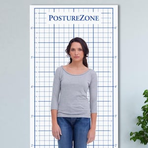May include: A woman stands in front of a white grid with blue lines and the text "POSTUREZONE". The grid has measurements in feet and inches. She wears a gray long-sleeve shirt and blue jeans.