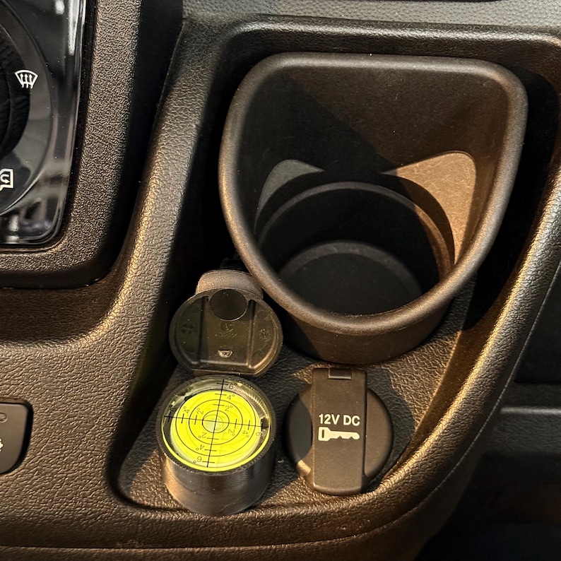 May include: A black car console with a cup holder, a 12V DC power outlet, and a round level with a yellow center. The level has a bubble and markings for precise measurements. The console is made of textured black plastic.