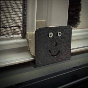Könnte beinhalten: Ein schwarzes, strukturiertes, quadratisches Objekt mit einem lächelnden Gesichtsdesign, das an einem Fensterrahmen befestigt ist. Das Gesicht wird mit einer kreisförmigen Nase und einem gebogenen Mund gestaltet. Zwei Schrauben dienen als Augen.
