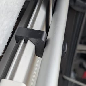 Könnte beinhalten: Nahaufnahme einer schwarzen Kunststoffhalterung, die eine silberne Metallstange sichert. Die Halterung ist an einer weißen Oberfläche befestigt. Die Stange hat ein kleines schwarzes Kunststoffteil.
