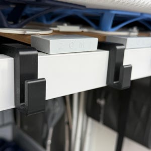 Könnte beinhalten: Nahaufnahme eines weißen Regals mit schwarzen Metallhaken. Hellgraue rechteckige Blöcke mit dem Wort "ZOMI" liegen auf dem Regal. Der Hintergrund zeigt weitere Regale und Gegenstände.