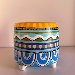 Op de afbeelding: Een cilindrische, handbeschilderde keramische plantenbak met een levendig, geometrisch ontwerp. De plantenbak heeft een gele rand, blauwe, groene en witte banden en een blauwe basis met witte accenten. Hij staat op drie kleine witte voetjes.